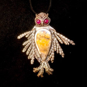May include: Silver owl pendant with red gemstone eyes and a teardrop-shaped yellow and brown stone center. The pendant has detailed wing and feather designs, suspended on a black cord. A unique piece of jewelry.