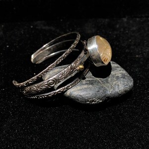 May include: A silver cuff bracelet with intricate floral and swirl patterns. A large, round, light-colored stone is set in a silver bezel and attached to the cuff. The bracelet is resting on a dark gray stone against a black background.
