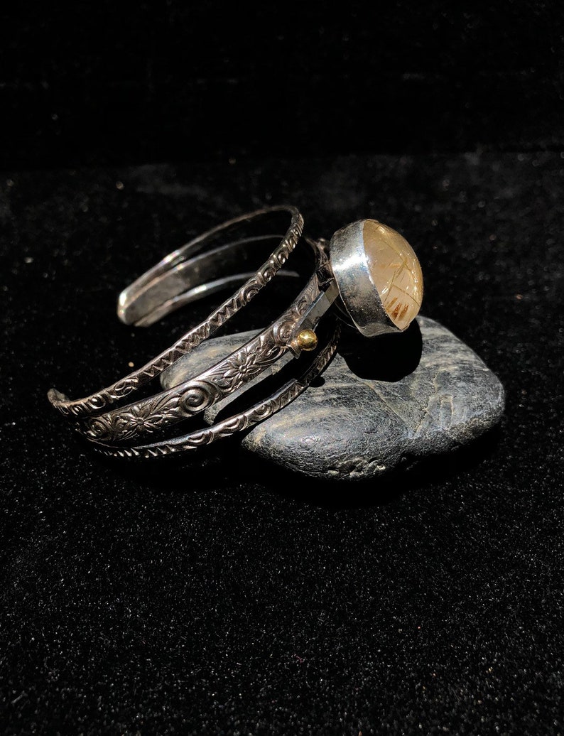 May include: A silver cuff bracelet with intricate floral and swirl patterns. A large, round, light-colored stone is set in a silver bezel and attached to the cuff. The bracelet is resting on a dark gray stone against a black background.