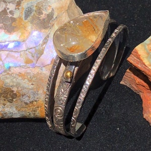May include: A silver cuff bracelet with a teardrop-shaped, clear quartz stone with golden inclusions. The bracelet features three bands with etched floral and swirl patterns, and a small gold accent.