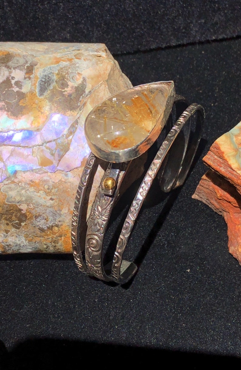 May include: A silver cuff bracelet with a teardrop-shaped, clear quartz stone with golden inclusions. The bracelet features three bands with etched floral and swirl patterns, and a small gold accent.