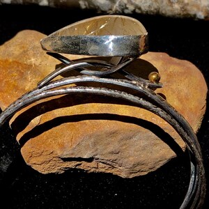 May include: A silver cuff bracelet with a large, teardrop-shaped, amber-colored stone. The stone is set in a silver bezel and sits atop a decorative silver setting. A small gold bead accents the design.