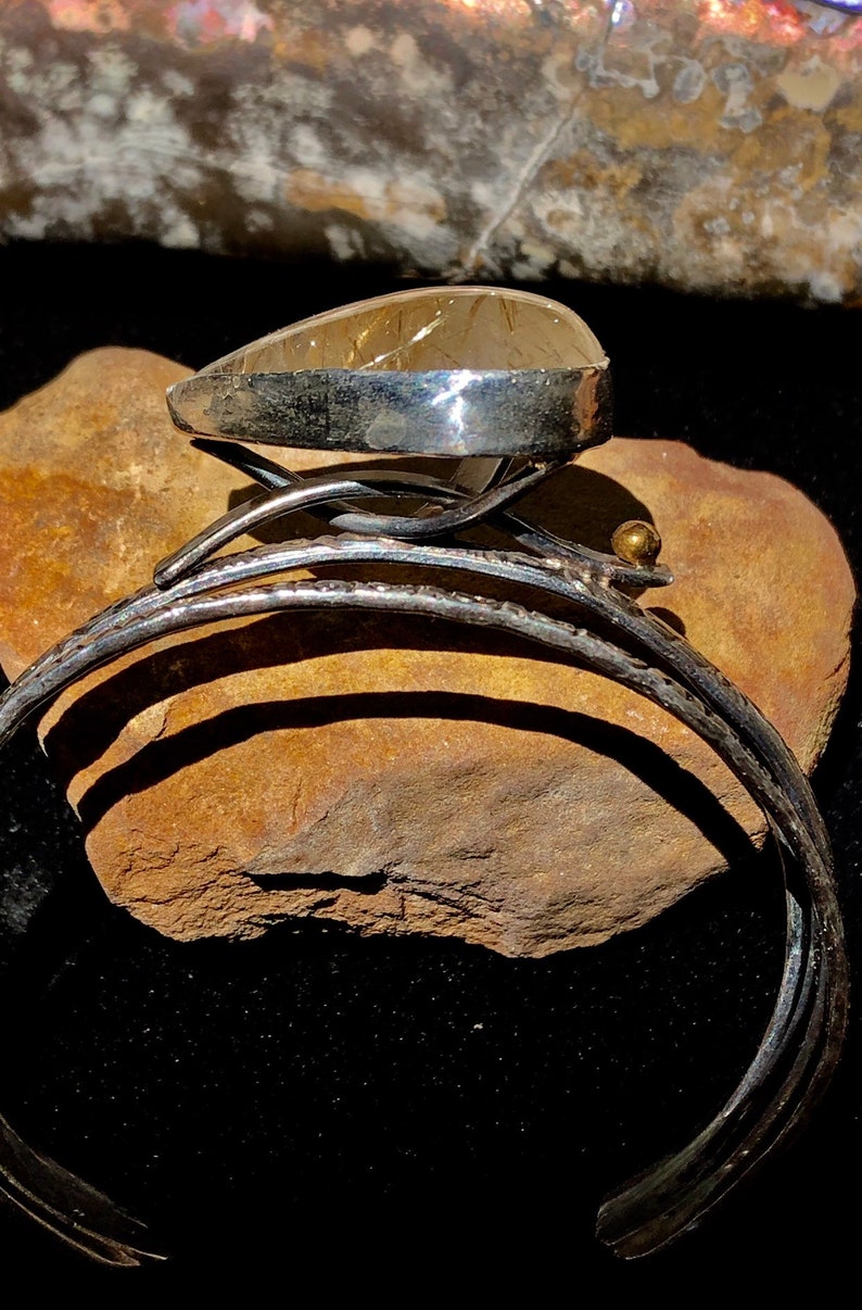 May include: A silver cuff bracelet with a large, teardrop-shaped, amber-colored stone. The stone is set in a silver bezel and sits atop a decorative silver setting. A small gold bead accents the design.