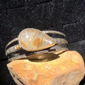 May include: A silver cuff bracelet with a teardrop-shaped, clear gemstone with gold inclusions. The bracelet has three bands, with engraved and textured details. The gemstone is set in a silver bezel. The bracelet is displayed on a brown rock against a black background.