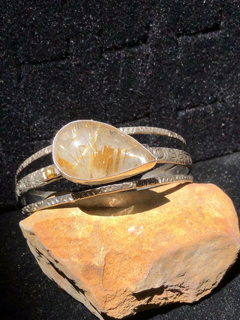 May include: A silver cuff bracelet with a teardrop-shaped, clear gemstone with gold inclusions. The bracelet has three bands, with engraved and textured details. The gemstone is set in a silver bezel. The bracelet is displayed on a brown rock against a black background.