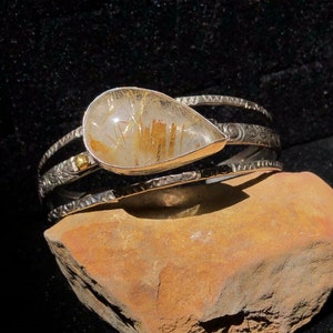 May include: A silver cuff bracelet with a teardrop-shaped, clear quartz stone containing golden rutile inclusions. The bracelet has intricate etched designs and a small gold accent. The jewelry is displayed on a brown stone against a black background.