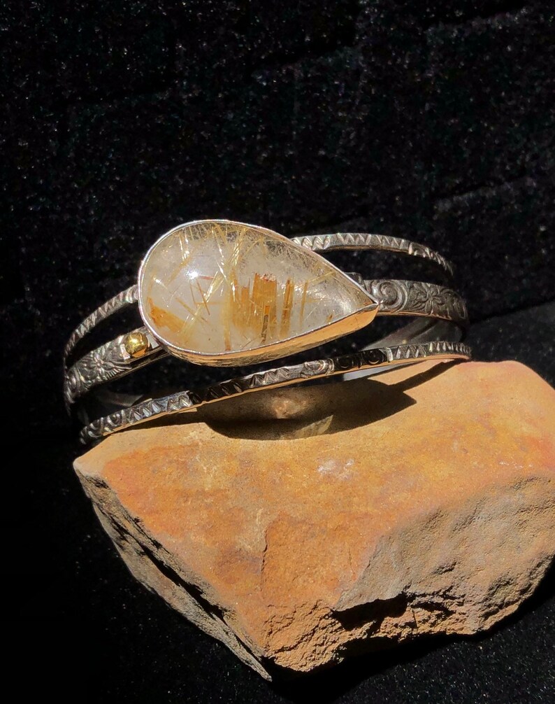 May include: A silver cuff bracelet with a teardrop-shaped, clear quartz stone containing golden rutile inclusions. The bracelet has intricate etched designs and a small gold accent. The jewelry is displayed on a brown stone against a black background.