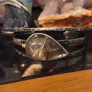 May include: A silver cuff bracelet with a large, teardrop-shaped, clear quartz stone with golden inclusions. The bracelet has an etched design and is displayed on a reflective surface. Other crystals are visible in the background.
