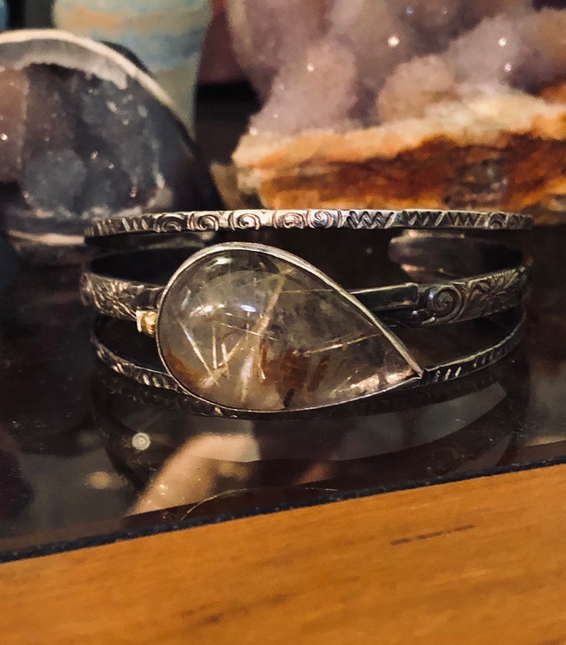 May include: A silver cuff bracelet with a large, teardrop-shaped, clear quartz stone with golden inclusions. The bracelet has an etched design and is displayed on a reflective surface. Other crystals are visible in the background.