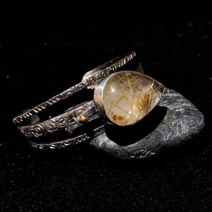 May include: A silver cuff bracelet with intricate floral and scrollwork designs. The bracelet features a large, teardrop-shaped gemstone with golden inclusions. The jewelry is displayed on a dark, textured surface, highlighting the details.