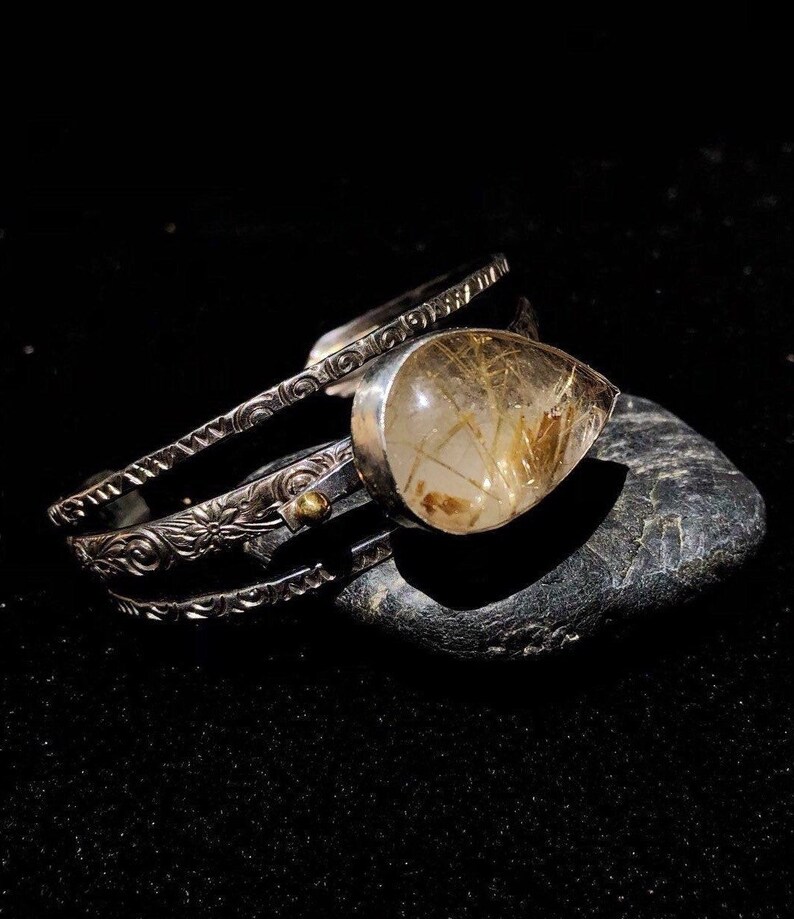 May include: A silver cuff bracelet with intricate floral and scrollwork designs. The bracelet features a large, teardrop-shaped gemstone with golden inclusions. The jewelry is displayed on a dark, textured surface, highlighting the details.