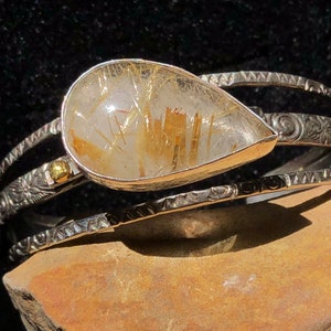 May include: A silver bracelet with a teardrop-shaped, clear quartz stone containing golden rutile needles. The bracelet has three bands with etched designs and a small gold accent. The jewelry is displayed on a brown stone surface.