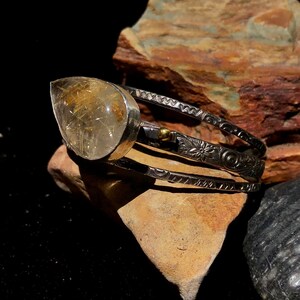 May include: A silver cuff bracelet featuring a large, teardrop-shaped, clear quartz stone with golden inclusions. The bracelet has three bands with etched floral and geometric patterns. The stone is set in a silver bezel.