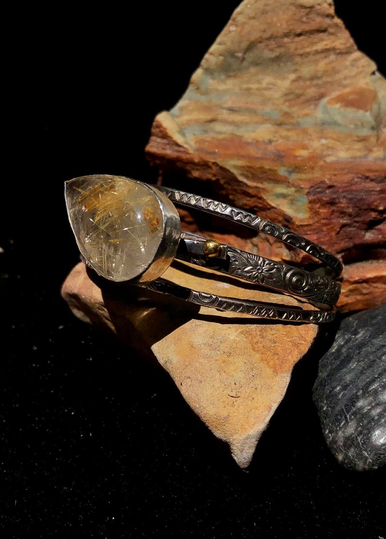May include: A silver cuff bracelet featuring a large, teardrop-shaped, clear quartz stone with golden inclusions. The bracelet has three bands with etched floral and geometric patterns. The stone is set in a silver bezel.
