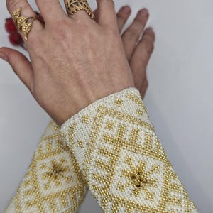 May include: Ivory-colored beaded wrist cuffs with a geometric gold pattern. The cuffs are adorned with small, round gold beads. Two gold leaf-shaped rings are visible on the fingers.