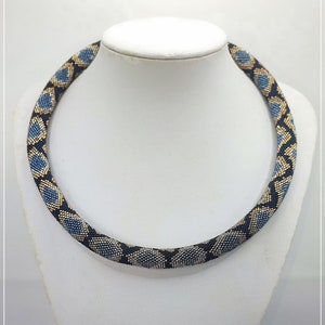 Puede incluir: Un collar de cuentas azul, dorado y negro con un patrón de piel de serpiente. El collar está en un maniquí blanco.