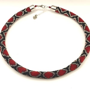 Puede incluir: Un collar de cuentas rojas, negras y plateadas con un cierre en forma de corazón. El collar está hecho de pequeñas cuentas que se tejen juntas para crear un patrón geométrico.