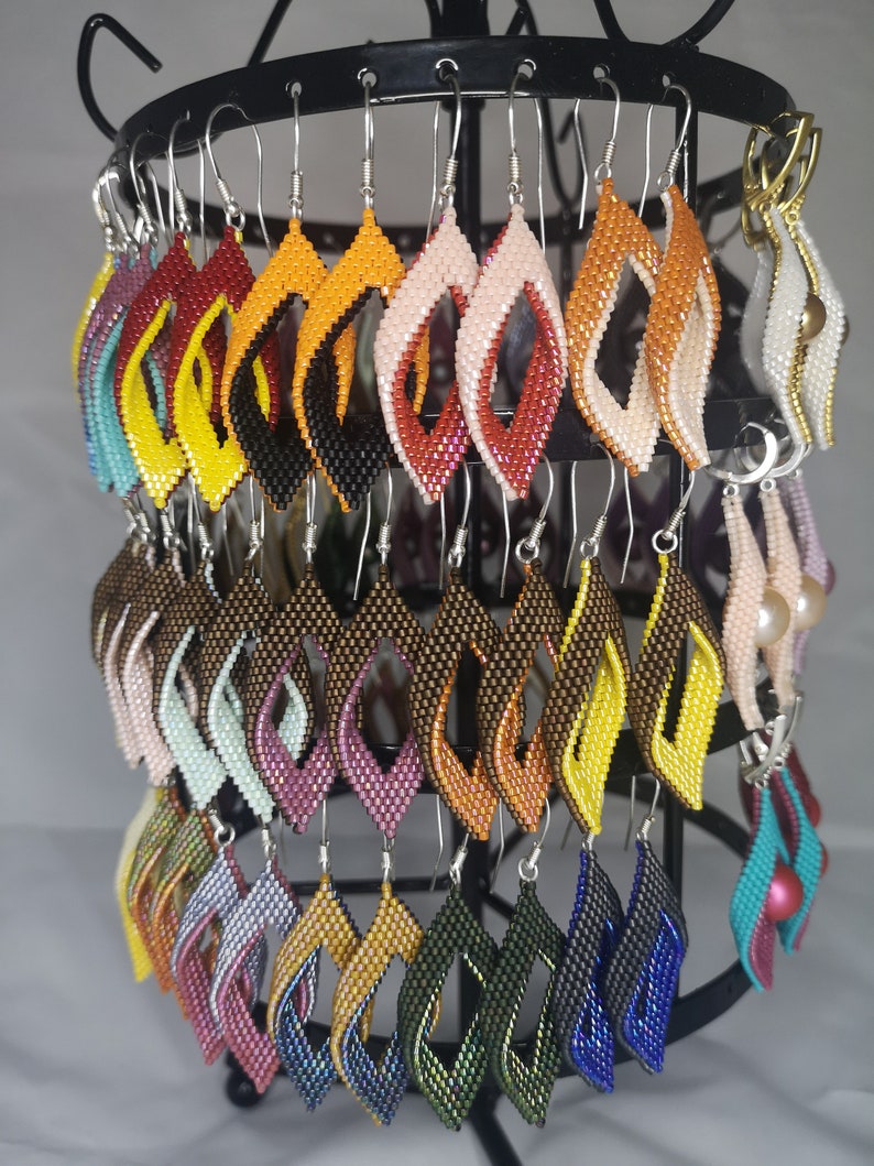 May include: A display of colorful beaded earrings in various shades of red, orange, yellow, green, blue, purple, and brown. The earrings are displayed on a black metal stand with hooks.