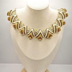 May include: A beaded necklace with a geometric design. The necklace features alternating sections of white, brown, and gold beads, creating a zig-zag pattern. The necklace is displayed on a mannequin bust.