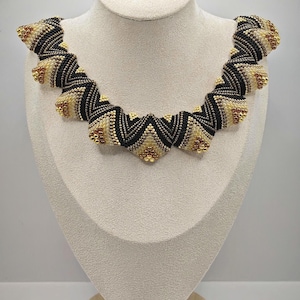 May include: A beaded necklace displayed on a mannequin. The necklace features a repeating pattern of black and gold beads, creating a zigzag design. The necklace is a statement piece, perfect for adding a touch of elegance to any outfit.