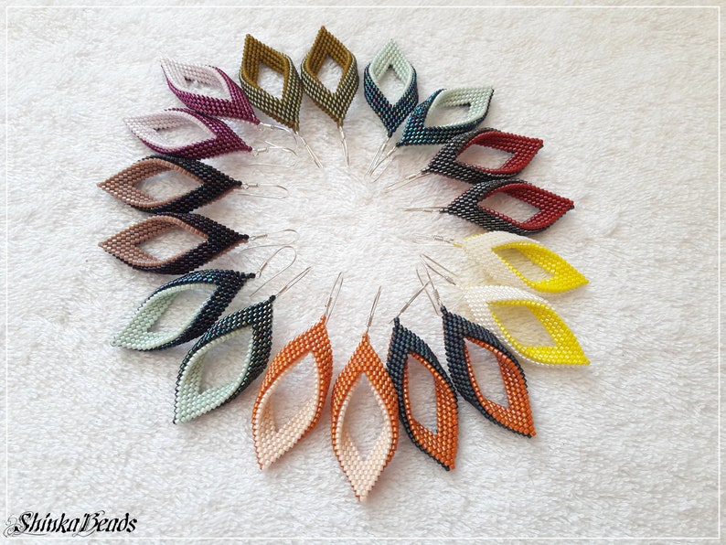 May include: A collection of 12 pairs of beaded earrings in various colors, including black, white, red, orange, yellow, green, and brown. The earrings are arranged in a circle on a white surface. Each earring is made of two overlapping teardrop shapes, creating a unique and stylish design.