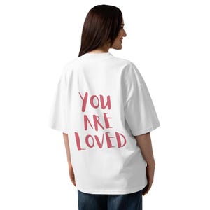 May include: A white oversized t-shirt with the words "You Are Loved" printed in a pinkish-red color on the back. The shirt has short sleeves and a relaxed fit. The model is wearing blue jeans.