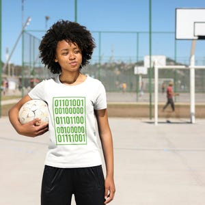 Pode incluir: Camiseta branca com código binário verde. A pessoa está segurando uma bola de futebol. O fundo inclui uma cesta de basquete e uma baliza de futebol em um dia ensolarado.