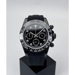 Könnte beinhalten: Eine schwarze Armbanduhr mit schwarzem Zifferblatt und schwarzem Kautschukarmband. Die Uhr hat drei Hilfszifferblätter und Diamant-Stundenmarkierungen. Die Lünette hat weiße Markierungen und den Text "UNITS PER HOUR". Die Uhr wird auf einem schwarzen Ständer präsentiert.