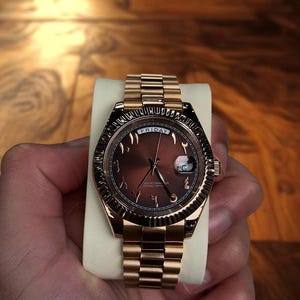 Puede incluir: Un reloj de pulsera dorado con esfera marrón y números arábigos. El reloj presenta un bisel estriado y un brazalete dorado a juego. La palabra "FRIDAY" se muestra en la parte superior de la esfera.