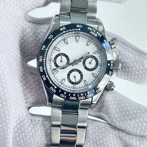 May include: A stainless steel wristwatch with a white face and black bezel. The watch features three sub-dials, a date display, and a silver-tone bracelet. The bezel has the text "UNITS PER HOUR".