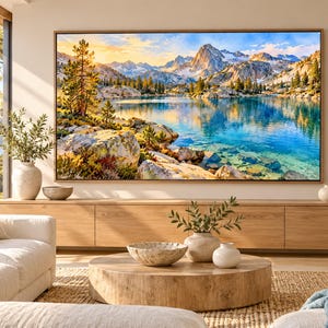 May include: A large rectangular wall art print featuring a vibrant landscape of a clear blue mountain lake, rocky shores, and evergreen trees under a golden sky, displayed in a thin wooden frame above a long light wood media console.