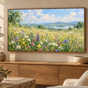 May include: A rectangular landscape painting in a thin wooden frame depicting a vibrant meadow filled with white, yellow, purple, and pink wildflowers under a bright blue sky with white clouds, overlooking rolling green hills and a distant lake.