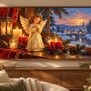 Puede incluir: Un cuadro rectangular de pared que muestra la pintura de un &aacute;ngel con alas doradas y las manos juntas en oraci&oacute;n. La escena incluye velas rojas y blancas, ramas de pino y un farol, frente a un paisaje de pueblo invernal nevado.