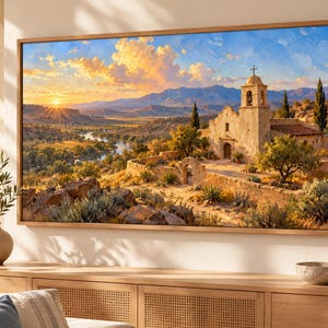 May include: A rectangular framed wall art print depicting a landscape with a stone mission church, a winding river, and distant mountains under a golden sunset sky. The scene features warm orange and yellow light, green vegetation, and rocky terrain.
