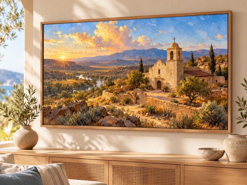 May include: A rectangular framed wall art print depicting a landscape with a stone mission church, a winding river, and distant mountains under a golden sunset sky. The scene features warm orange and yellow light, green vegetation, and rocky terrain.