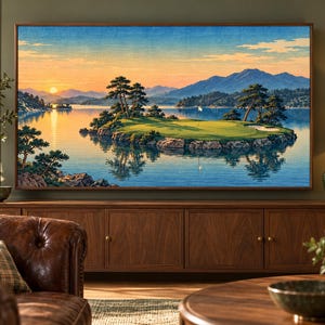 Puede incluir: Una impresi&oacute;n de paisaje enmarcada que representa un green de golf en una peque&ntilde;a isla rodeada de agua, con pinos, monta&ntilde;as lejanas y un cielo al atardecer en tonos naranja, amarillo y azul, exhibida en una pared sobre un mueble.