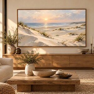 May include: A large rectangular landscape painting in a thin wooden frame depicting a coastal scene with sand dunes, beach grass, and a sunset over the ocean. The artwork is mounted on a wall above a long wooden media console in a living room.