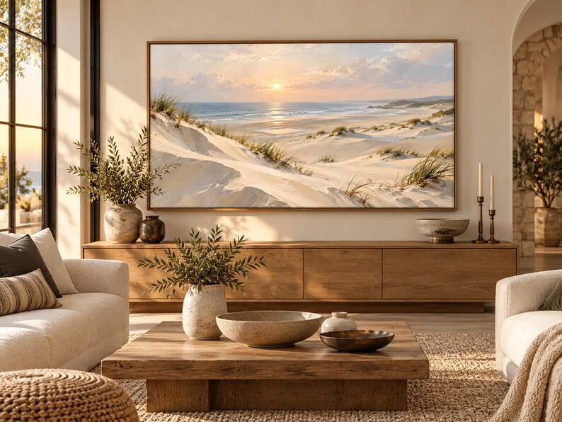 May include: A large rectangular landscape painting in a thin wooden frame depicting a coastal scene with sand dunes, beach grass, and a sunset over the ocean. The artwork is mounted on a wall above a long wooden media console in a living room.