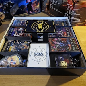 Puede incluir: Una caja negra que contiene un juego de cartas de League of Legends. El juego incluye cartas, dados y figuras de personajes. La caja tiene el logotipo de League of Legends en la parte delantera y en una tarjeta grande.