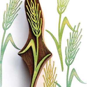 Può includere: Un timbro in legno marrone scuro con un design intagliato a forma di stelo di grano. Il timbro è posizionato su una superficie bianca che mostra diverse impronte di steli di grano verdi e gialle, che illustrano l'impressione del motivo.