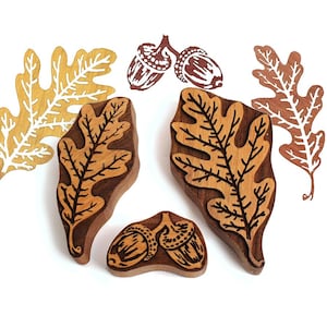 May include: Three wooden stamps featuring carved designs of two oak leaves and a pair of acorns. The stamps are accompanied by two printed paper cutouts of oak leaves and a printed outline of two acorns, all displaying natural wood tones.