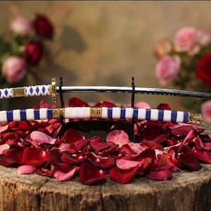 May include: Two decorative miniature katana swords with white and blue handles, displayed on a black stand atop a bed of red rose petals. The swords have gold accents and are set against a blurred floral background.