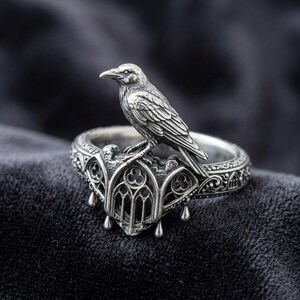 May include: A silver-colored ring featuring a detailed raven perched atop an ornate, gothic-style setting. The ring has a detailed design with three teardrop-shaped pendants. The ring is set against a dark background.