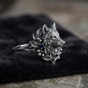 May include: A silver ring featuring a detailed wolf head sculpture surrounded by sculpted leaves and small berries. A single small, round, dark blue gemstone is set among the foliage. The band of the ring is shaped like a textured tree branch.