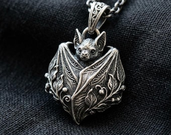 May include: A silver pendant featuring a detailed bat with its wings wrapped around its body, adorned with intricate leaf and vine carvings. The pendant hangs from a matching silver chain, showcasing a dark, oxidized finish that highlights the texture.