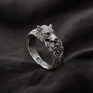 Peut inclure: Bague en argent ornée d'une tête de loup détaillée. La bague présente un motif tourbillonnant orné sur le bracelet et comporte une pierre noire et deux petites pierres violettes. La bague est posée sur un fond sombre.