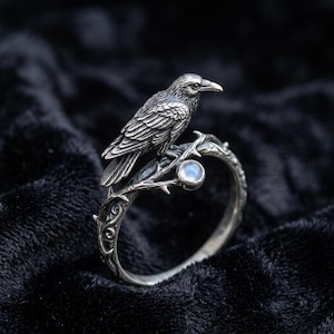 May include: A silver-toned ring featuring a detailed raven perched on a branch with a small, round, light blue gemstone. The ring's band has an intricate, textured design, and the raven's feathers are finely detailed.