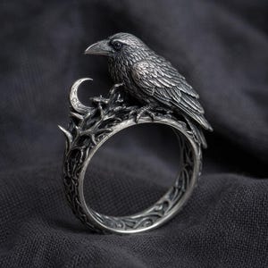 May include: A detailed silver ring featuring a raven perched on a branch with a crescent moon. The ring has an intricate design with a dark, antique finish. The raven's feathers and the branch's texture are finely detailed.