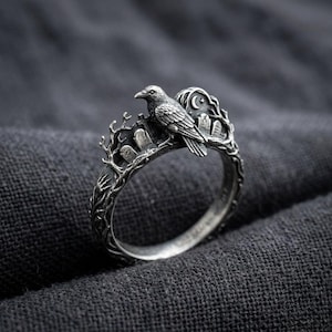 May include: A silver-toned ring featuring a detailed raven perched on a branch. The band incorporates a graveyard scene with upright tombstones, bare tree branches, and a small crescent moon with a star set against a dark, textured metal finish.