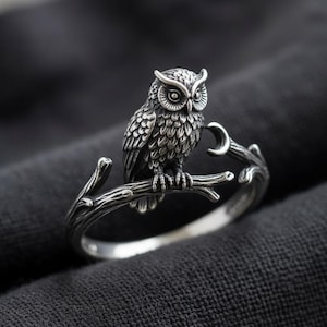 Könnte beinhalten: Ein silberfarbener Ring mit einer detaillierten Eule, die auf einem strukturierten Ast sitzt. Die Ringschiene ist wie ein Baumzweig geformt, und eine kleine Mondsichel ist in das Design nahe der Eule integriert, mit einem oxidierten Finish.